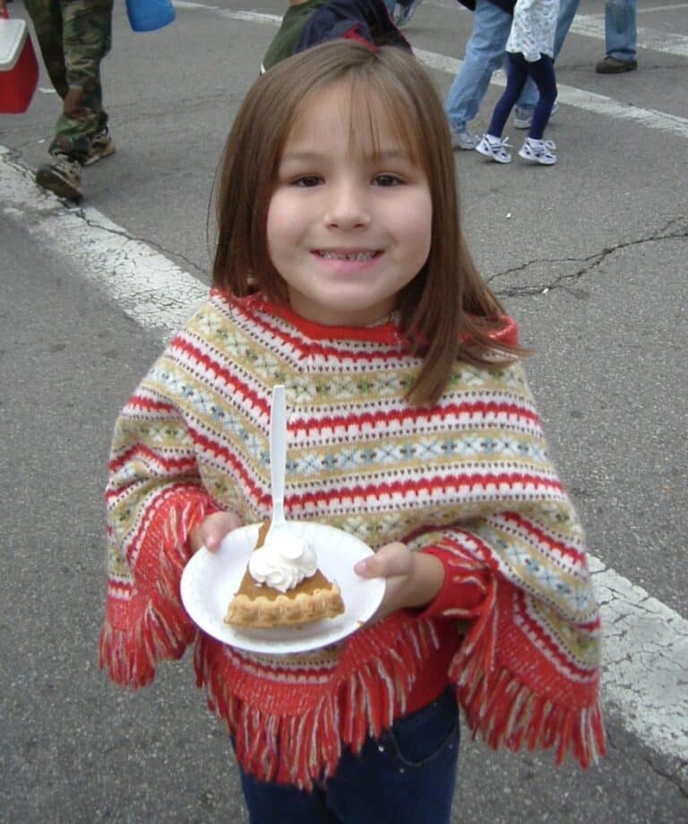 Little girl wearing a red poncho, holding a piece of pumpkin pie with whipped cream on top.  