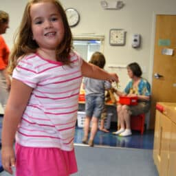 A young Caucasian girl in a pink and white striped shirt and pink skirt. She's in a classroom.